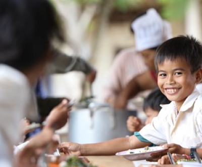 Boy smiles at the camera, surrounded by his schoolmates eating their free school meal in Cambodia