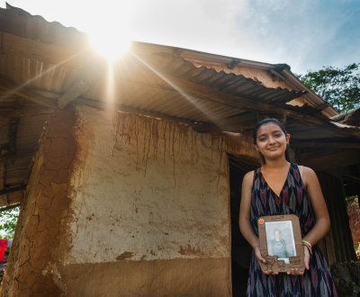 Sponsored girl from Honduras standing outside her house and holding up a portrait photo of her sponsor.