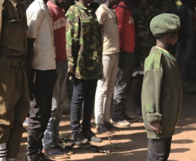 People stand in line behind a child wearing army camouflage and a beret in South Sudan