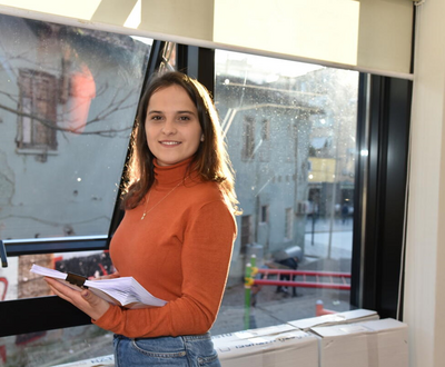 Girl from Albania standing next to a window and holding a book while smiling to the camera