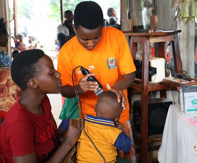 Girl in Tanzania shaving a child's head while his mother holds him.