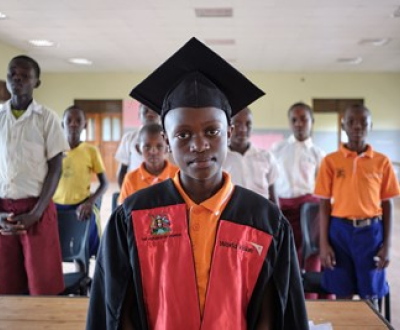 Boy from Uganda standing in a classroom in front of other boys, facing the camera while wearing a graduation hat