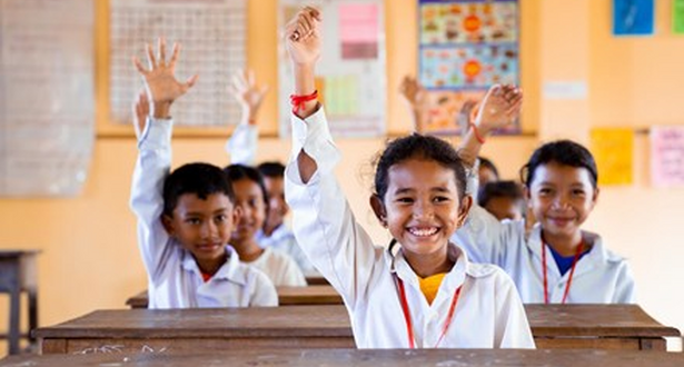 A class of children from Cambodia pictured wearing school uniforms in a classroom and all raising their hand and smiling