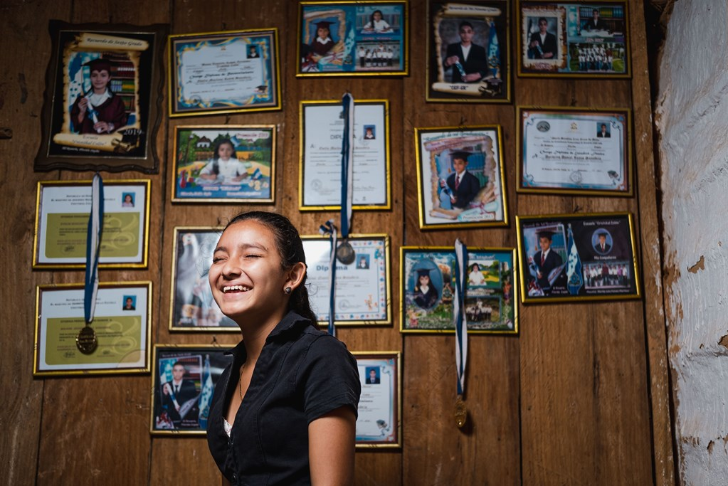 Girl from Honduras laughing while standing in front of a wall with certificates and ribbons hung on it