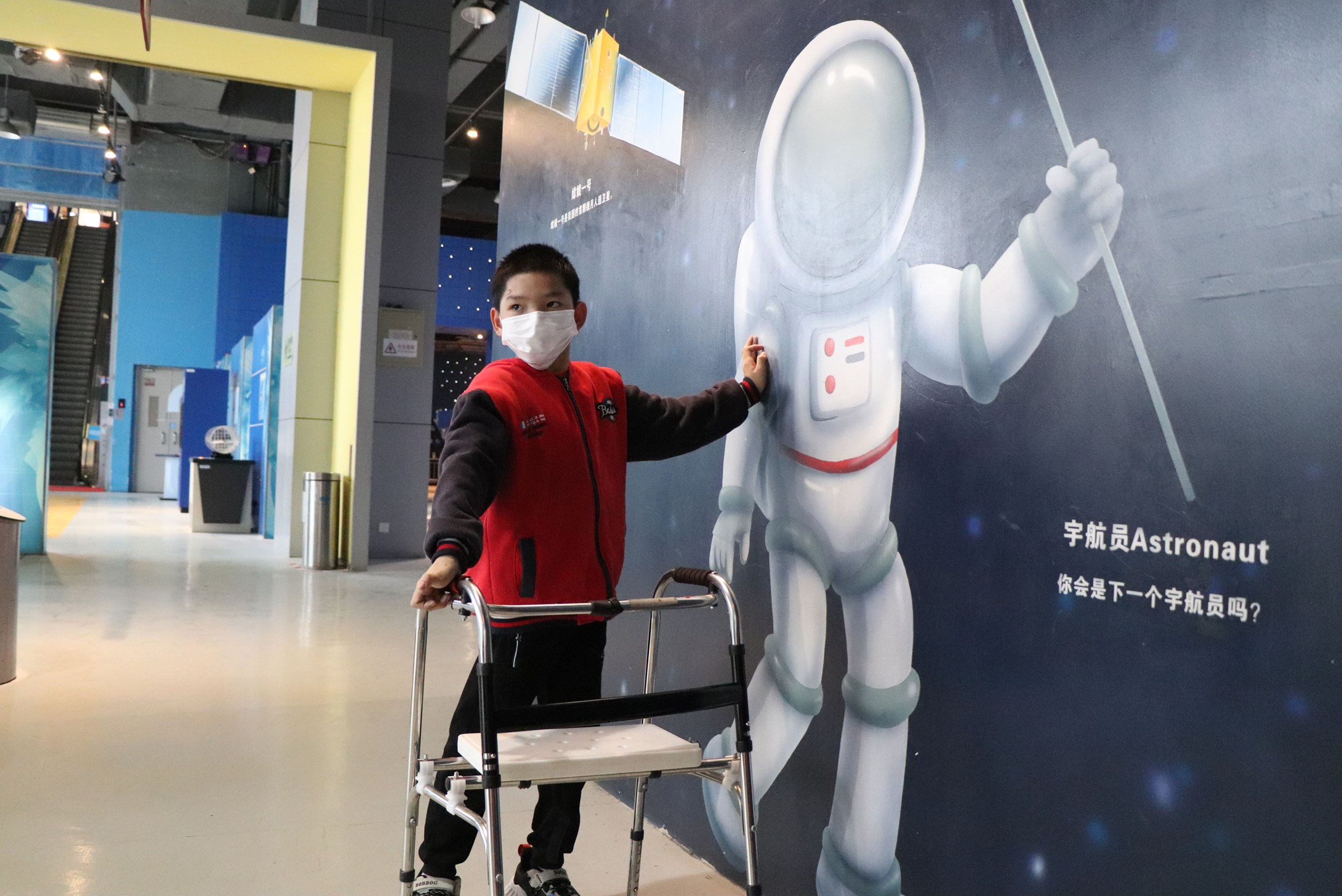 Disabled child from China wearing a facemask and standing next to a poster of an austronaut.